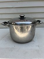 Stainless steel stockpot with glass lid and metal knob seen from front and side angle on neutral textile surface.