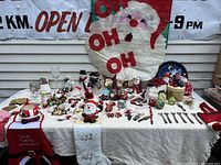 Overview of the lot on a white tablecloth with many Santa Claus figurines, stockings, ornaments including candy canes, and a large 'OH OH OH' Santa face wall decoration hanging above.