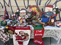 Photo showing a broad assortment of Christmas decorations including the colorful puffy wreath, various Santa figurines, holiday textiles with Christmas messages, snowmen, rocking chair decoration, and small ornaments on a table.
