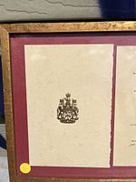 Framed item front showing cream paper with Canadian Coat of Arms emblem on dark red matte and gold-tone frame.