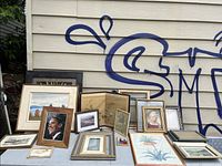 Multiple framed art pieces displayed outdoors showing a variety of styles including portraits and prints in different wood and metallic frames.