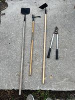 Four garden tools lying on pavement: hoe, digging tool, mattock, and pruning shears