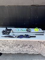 Full view of lot items including 3 golf drivers with covers, golf balls in packs and nets, and folded Wind River golf umbrella on table with lace tablecloth.