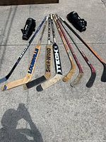 Photo showing nine used hockey sticks and two black goalie leg pads on concrete pavement.