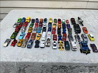 Overview of approximately 60 miniature toy cars arranged in rows on a lace-covered table showing a variety of colors and styles.