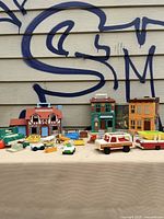 Overview showing three Fisher-Price dollhouses/buildings with assorted toy furniture and vehicles spread in front.
