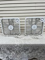 Two white Genesis brand room-sized box fans side by side on a patterned cloth surface outdoors.