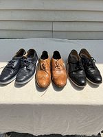 Three pairs of men's shoes displayed on a cloth surface outdoors under sunlight, showing front and side views.
