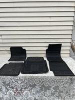 Five black rubber automotive floor mats arranged on a surface, showing varied sizes and patterns including some with grooves and textured surfaces.