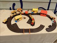 Overview of wooden train set on a beige cloth surface with brown plastic tracks arranged in a loop and wooden train cars assembled on track. Two yellow molded cabins and small wooden accessories including animals and trees visible.