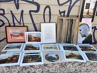 Table laid with multiple art prints and paintings including a framed landscape, a pen and ink drawing, abstract and modern portraits, arranged on a lace cloth.