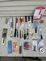 Top-down image showing assortment of new old stock kitchen knives and utensils laid on tablecloth, including peelers, scissors, tongs, timers, and knives with varying handle colors and packaging states.