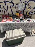 Full view of table with assorted nostalgic vintage kitchenware and decorative items, covered with lace tablecloth with outdoor storage tote in front