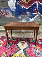 Wooden piano bench shown from side angle on a colorful rug, displaying its sturdy four legs and rectangular shape