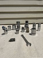 Group photo of 12 socket wrench accessories arranged on white textured surface, showing different socket sizes, two universal joints, and one short wrench