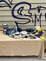 Photo of the lot items arranged on table including boxed Wahl Jumbo massage vibrator, yellow desk lamp, electronics kit, album cleaner, and small gadgets