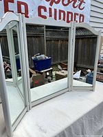 Image showing three connected mirror panels with white painted wood frames. The vanity mirror is freestanding and positioned outdoors with a visible fence and some items in the background.