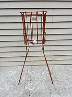 Red metal wire tennis ball hopper standing on four legs on a patterned floor with one white tennis ball inside.