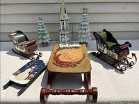 Overview of all tin Christmas decorations, including three sleds, two pine tree figurines, and one church steeple with 'Believe' sign on large sled.