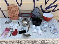 Photo of varied kitchen items arranged on a lace-covered table showing baking molds, cookware, tea set, utensils, and bowls.