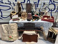 Wide shot of the lot showing a variety of wooden items including a small keyboard deck from a pump organ, wooden shoe stretchers, wooden hand plane, cedar shoe foot decoys, and other wooden tools and objects arranged on a table with a patterned cloth and a burlap sack underneath.