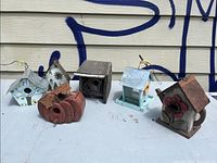 Front view of all six vintage birdhouses with background of siding and graffiti, placed on a white table in outdoor setting.