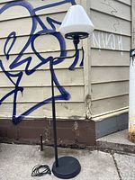 Full view of vintage IKEA Alvangen standing lamp with black metal base and milk glass shade, outdoors against beige siding with graffiti.