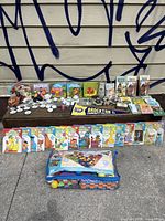 Wide view of vintage Ladybird books, other children's books, collectible toys, Brockton Community College pennant, flashcards, and plastic container with toys, laid out on a bench and ground.