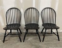 Front view of three black Windsor chairs with spindle backs, pegged legs, and sculpted seats, arranged side by side on a concrete floor against a plain background.