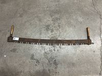 Antique two-person bow saw laid flat on concrete floor, showing full length with wooden handles and metal blade with teeth.