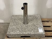 Granite square umbrella stand base with polished stainless steel central pole and adjustable knob, placed on a wooden pallet in an industrial environment.