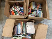 Wide view of three cardboard boxes filled with various vintage children's books showing colorful covers, text, and some boxed character items