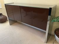 Front-left angled view of sideboard showing chrome sides, dark brown lacquer doors, and glass top.