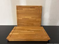 Two rectangular wooden cutting boards displayed, one standing vertically behind a larger board lying flat. Both show a block-pattern wood surface, larger board with a groove, smaller with a chipped corner.