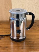 Front view of Nespresso Aeroccino milk frother showing stainless steel body, black handle, power button, and power cord on wooden surface.