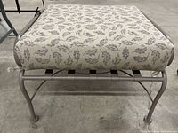Front angled view of the outdoor wrought iron ottoman stool showing the patterned cushion and metal base.