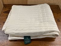 Folded Ralph Lauren cream cable knit throw blanket on a wooden surface, showing knit texture.