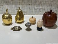 Front view of all vintage decorative pieces including gold-colored wood apple and pear, stone apple and egg, dark wood apple box, pewter mushroom, and brass turtle box.