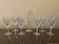 Seven crystal glasses arranged in two rows on a reflective dark surface against a beige wall, showing 6 liquor glasses and 1 champagne flute in the middle.