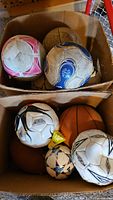Two cardboard boxes filled with assorted sports balls including various soccer balls, basketballs, a shuttlecock, and two footballs.