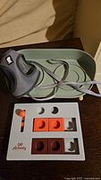 Photo showing gray cat harness with leash on a green feeding tray, and white Cat Activity puzzle toy with orange and brown sliders and orange knob