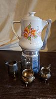Front view of 12 cup Italian espresso pot with a white porcelain top featuring floral designs. Silver plated creamer and sugar bowl included in the set shown in front.