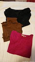 Three folded long sleeve tops in brown, fuchsia, and black with lace sleeves on a flat surface.