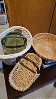 Lot of four wicker baskets displayed on a table in front of a wooden background. Visible are the large white wicker hamper basket holding the small green basket, the round natural wicker basket, and the rectangular beige basket with leather handles.