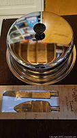 Stainless steel cake dish with lid, shiny reflective surface, black knob handle on top, photographed on wooden tabletop