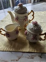 Photo of cream-colored tea set including creamer, sugar bowl, and coffee/tea pot with pink rose appliques and gold accents on a beige patterned cloth.