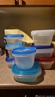 Stack of various plastic food storage containers with assorted blue, red, and white lids placed on a kitchen counter.