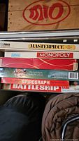 Partially stacked board games showing titles Masterpiece, Monopoly, and Battleship among others in box format with some wear.