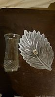 Clear view of the glass feather-shaped bowl and clear flower vase with textured base side by side on a dark surface