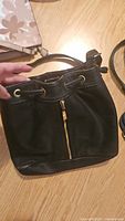 Black faux leather shoulder bag with a drawstring closure and a front vertical gold zipper pocket on a wooden surface, held by a hand.